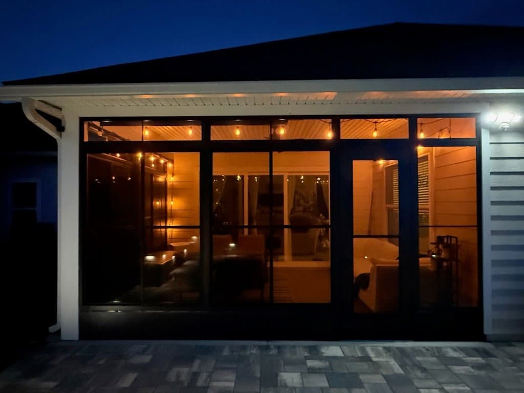 Enclosed porch glowing with string lights at night behind black-framed vinyl windows